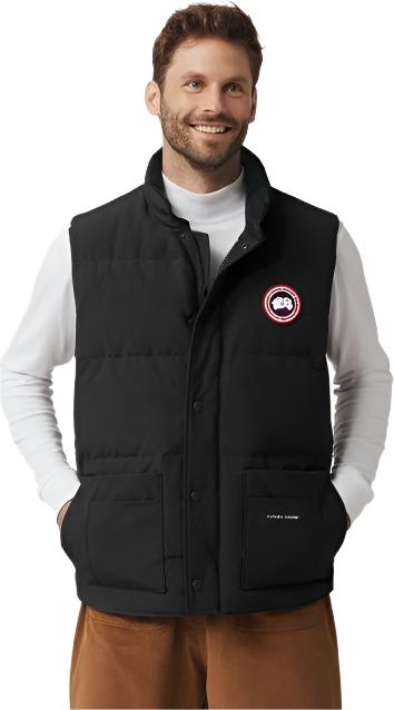 A man is wearing a black vest with a circular logo on the chest. He is wearing a white long sleeve shirt and brown pants.
