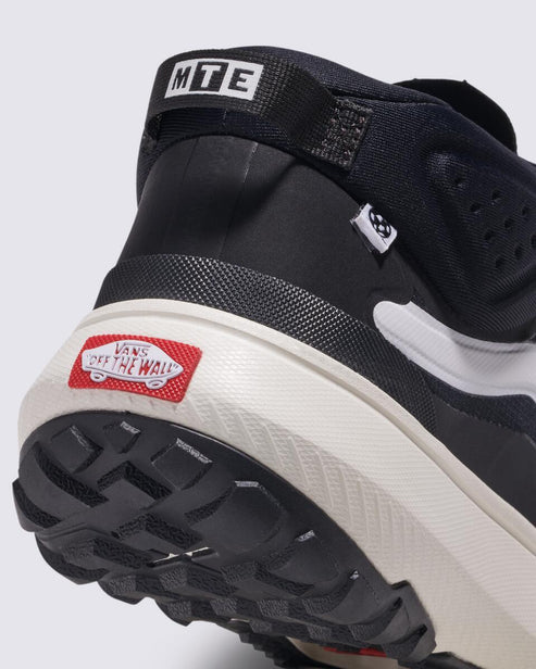 A close-up of the heel of a black and white shoe with a red 'VANS OFF THE WALL' logo on the cream-colored sole and 'MTE' stitched into the top of the black pull loop.
