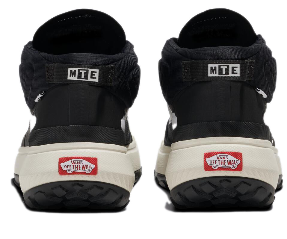 The rear view of a pair of black hi-top sneakers with a cream-colored sole. An MTE tag is above the heel and a Vans logo is at the heel. 