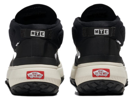 The rear view of a pair of black hi-top sneakers with a cream-colored sole. An MTE tag is above the heel and a Vans logo is at the heel. 