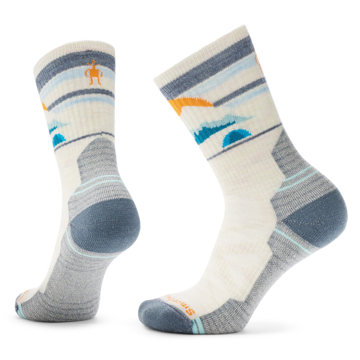 Two white crew socks with blue and orange mountain landscape designs, gray reinforced heel and toe, and small person-shaped logo on the ankle of one sock.