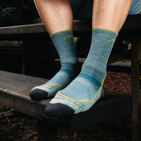 Person sitting on a wooden bench wearing teal crew socks with dark gray toes, heels and gold trim.