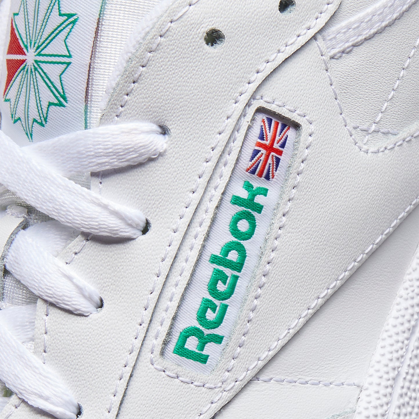 Close-up of a white shoe with white laces, a visible green logo with the word "Reebok" and a small British flag above the logo.