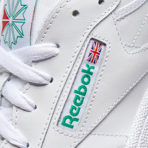 Close-up of a white shoe with white laces, a visible green logo with the word "Reebok" and a small British flag above the logo.
