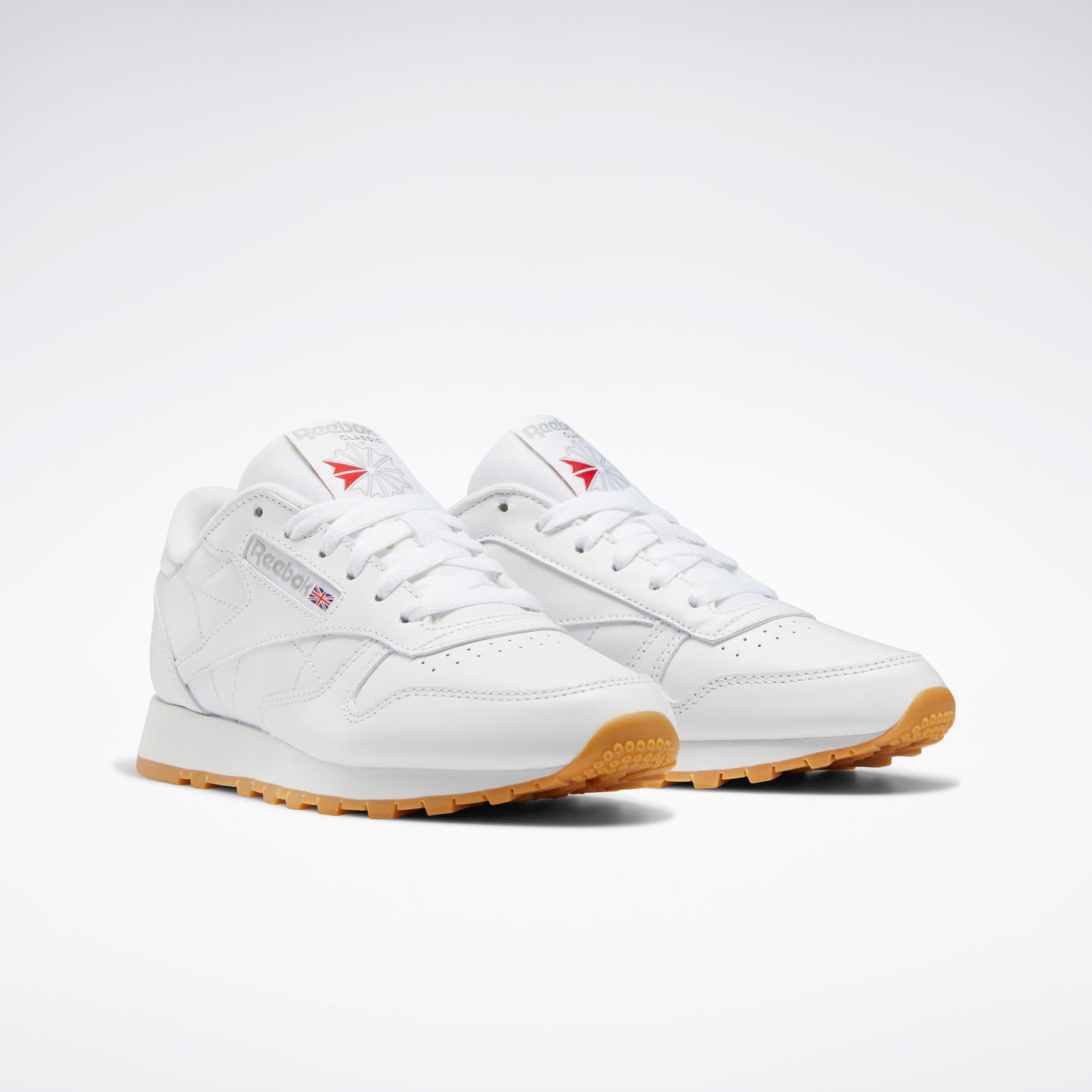 A pair of low-profile sneakers with white leather uppers, white laces, red logos on the tongue, and gum outsoles.