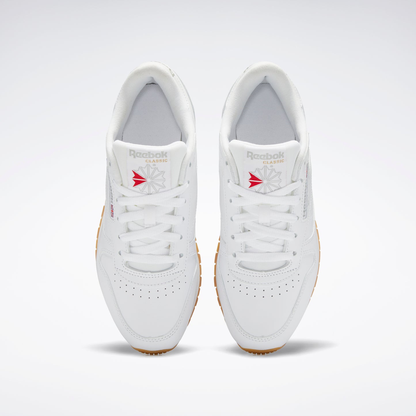 Top-down view of a pair of white sneakers with white laces and gum soles.  The tongue shows a logo with a red design.