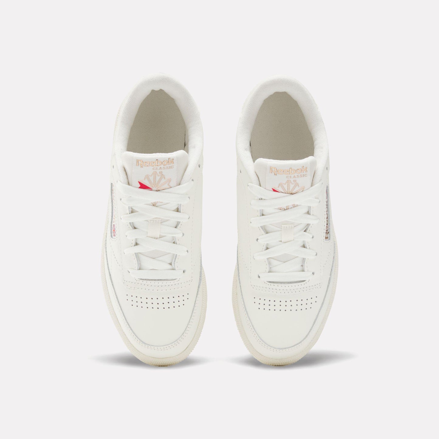 Top-down view of two white leather sneakers with white laces; logo tag on tongue with pink accents, and perforated toe box.