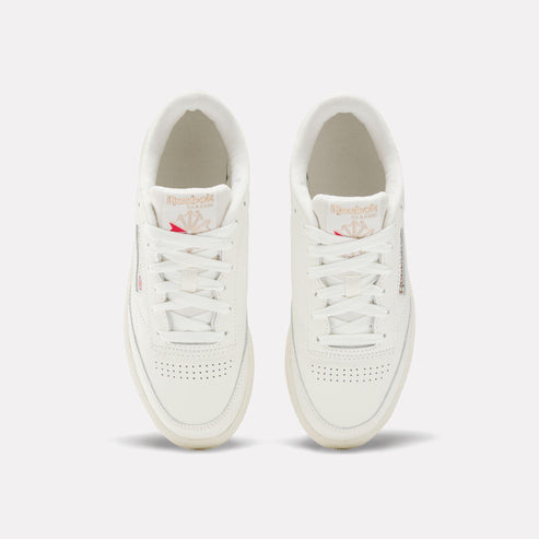 Top-down view of two white leather sneakers with white laces; logo tag on tongue with pink accents, and perforated toe box.