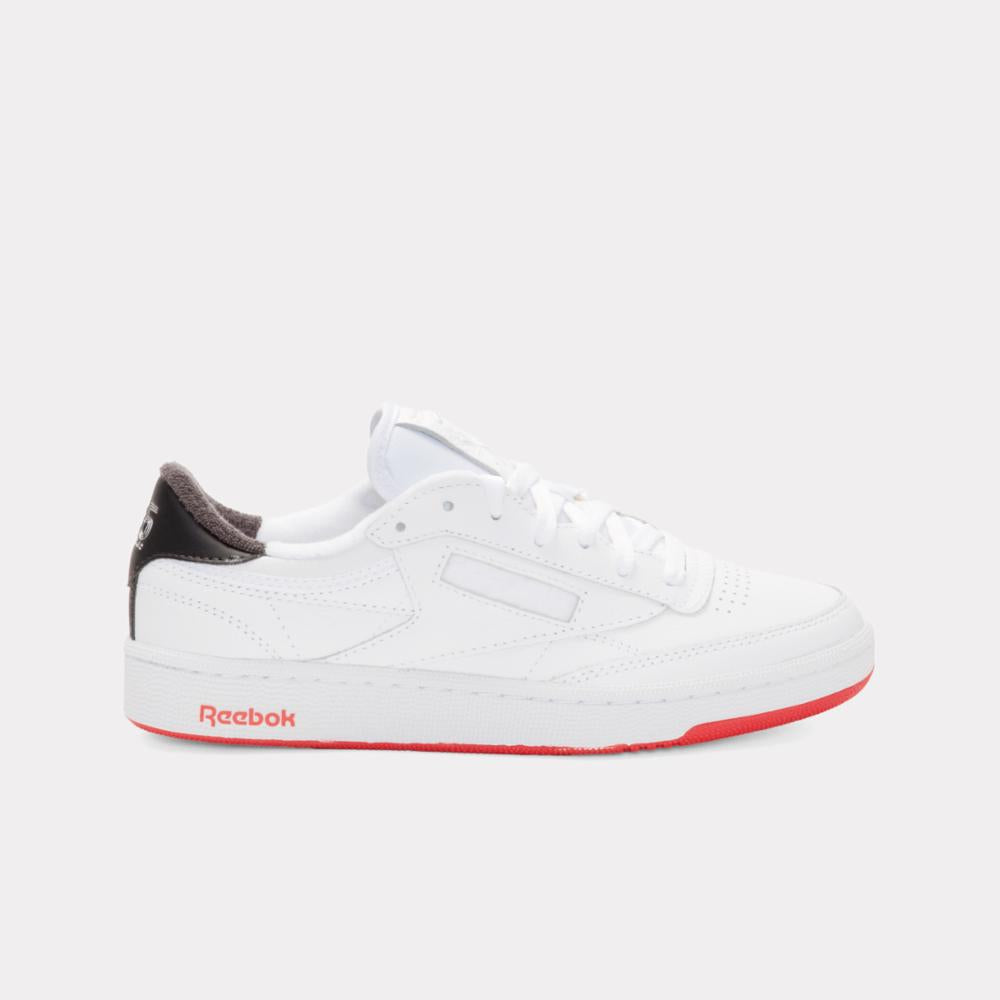 A white low-top sneaker with a black heel and a red sole with the word "Reebok" written on it.