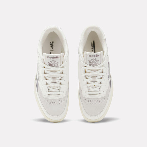 Two white sneakers facing forward, with white laces and a logo on the tongue.