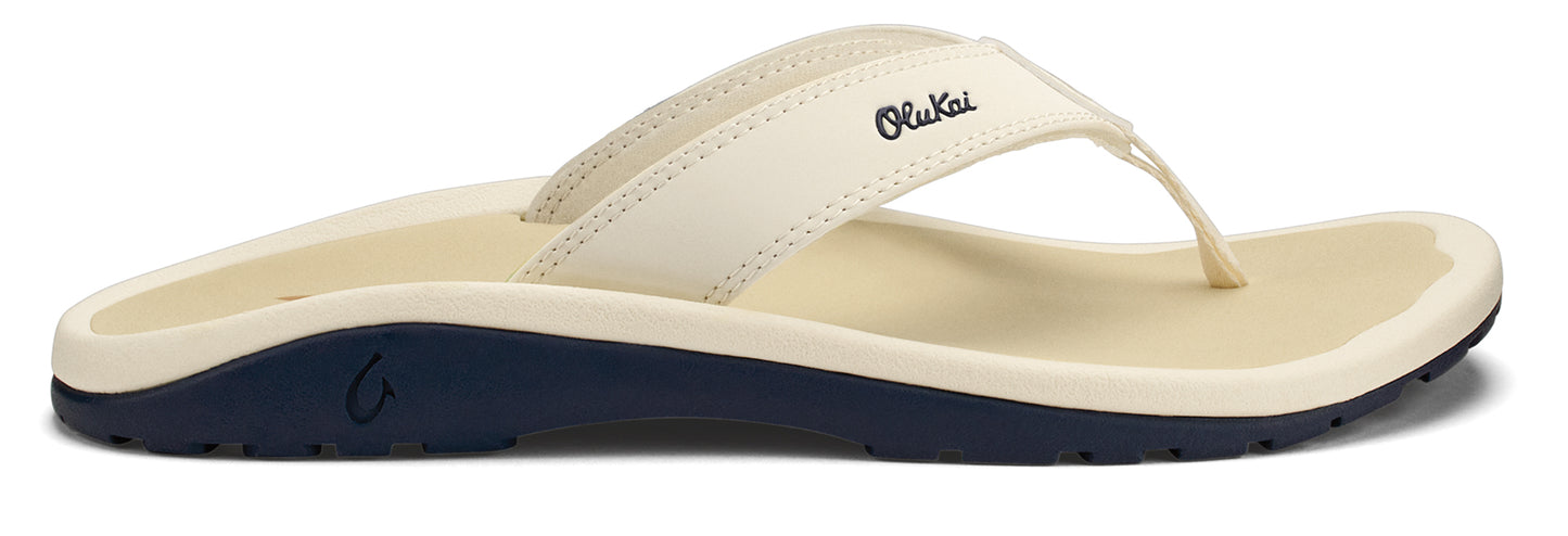 A flip-flop with light-colored straps and sole on a navy blue base; the brand name "OluKai" is visible on the side.