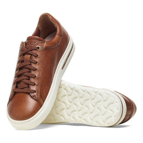 A pair of low cut light brown leather sneakers with white soles and brown laces, one sneaker on its side revealing the sole