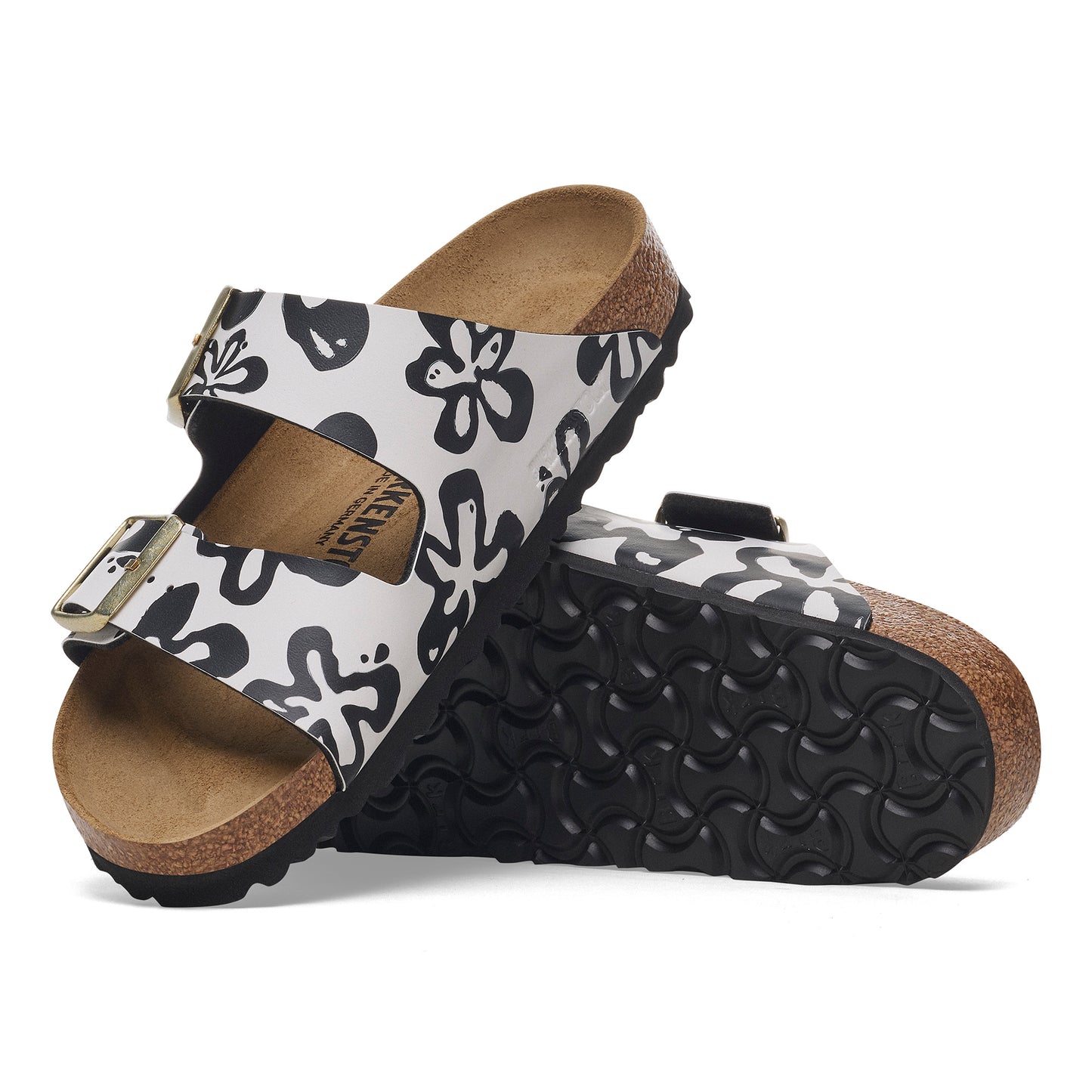 Two sandals with white floral patterned straps and dark soles are stacked on top of each other.