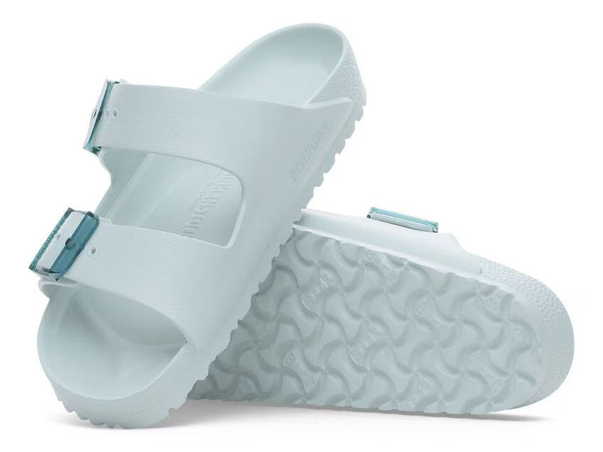 Two light-blue slide sandals with adjustable buckle straps, one resting on its side revealing the sole.