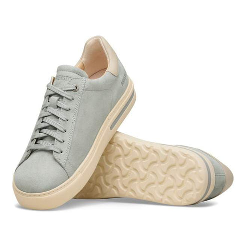 A pair of low-top sneakers with light gray suede uppers and off-white soles, one upright and one lying on its side revealing a wavy patterned outsole.