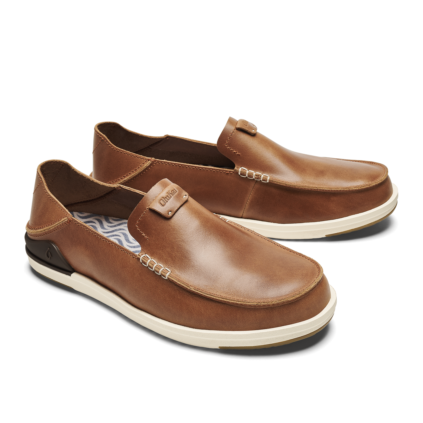 A pair of men's slip-on shoes made of brown leather with visible white stitching, an off-white rubber sole, and a dark heel accent, featuring a small tag with the text Olukai on the tongue.