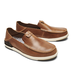 A pair of men's slip-on shoes made of brown leather with visible white stitching, an off-white rubber sole, and a dark heel accent, featuring a small tag with the text Olukai on the tongue.