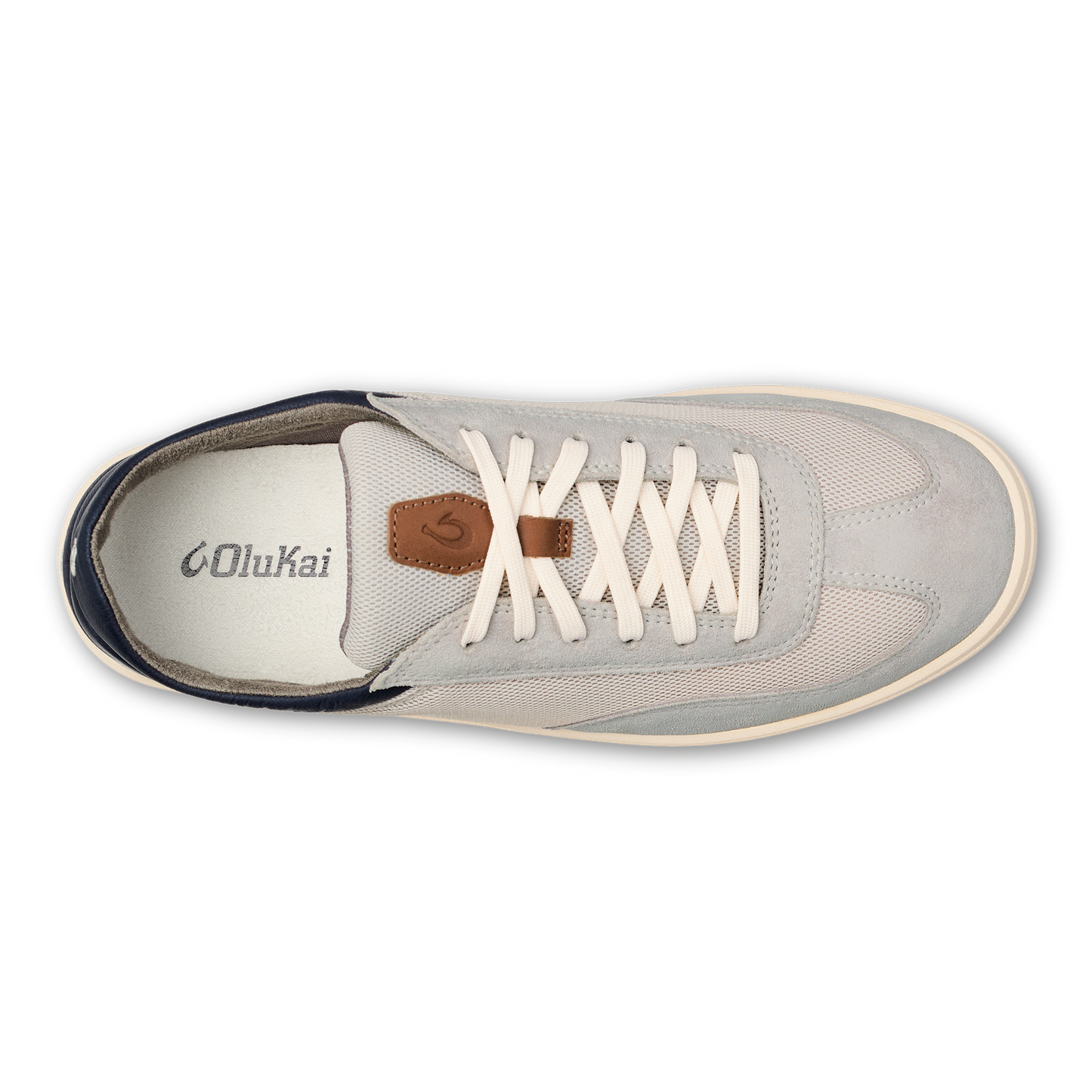 A light grey, top-down view of a slip-on sneaker with white laces, a tan leather accent on the tongue, a white insole with the OluKai logo, and a navy heel accent.