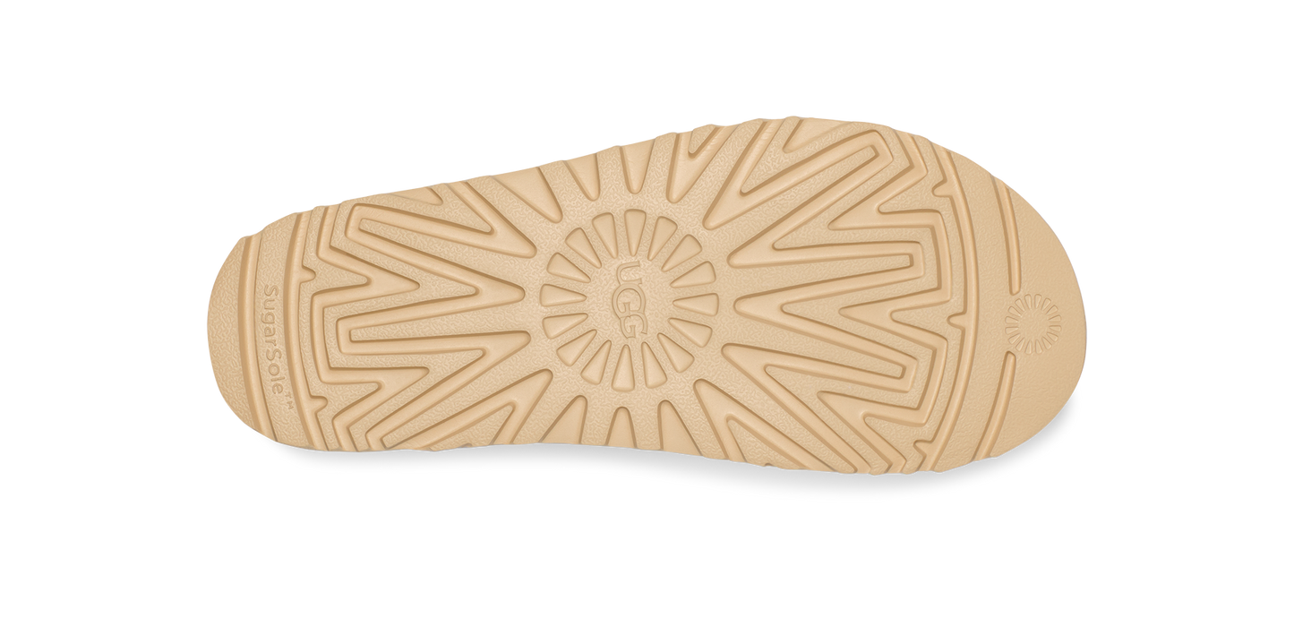 Close-up of the sole of a beige-colored shoe, featuring an intricate tread pattern with the text "UGG" embossed in the center.