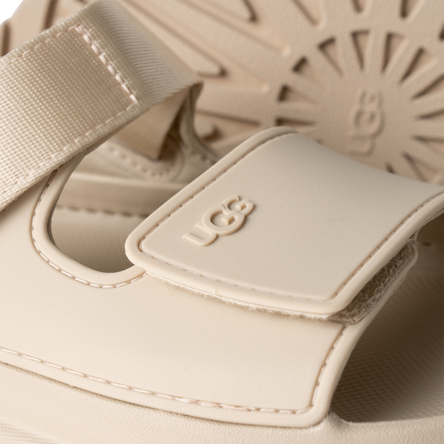 Close-up of a cream-colored sandal with the letters UGG embossed on the strap.