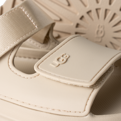 Close-up of a cream-colored sandal with the letters UGG embossed on the strap.