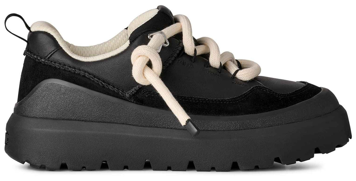 A black sneaker with a thick, rugged platform sole, featuring cream-colored rope laces, a black suede and synthetic leather upper, and a cream-colored lining at the heel collar.