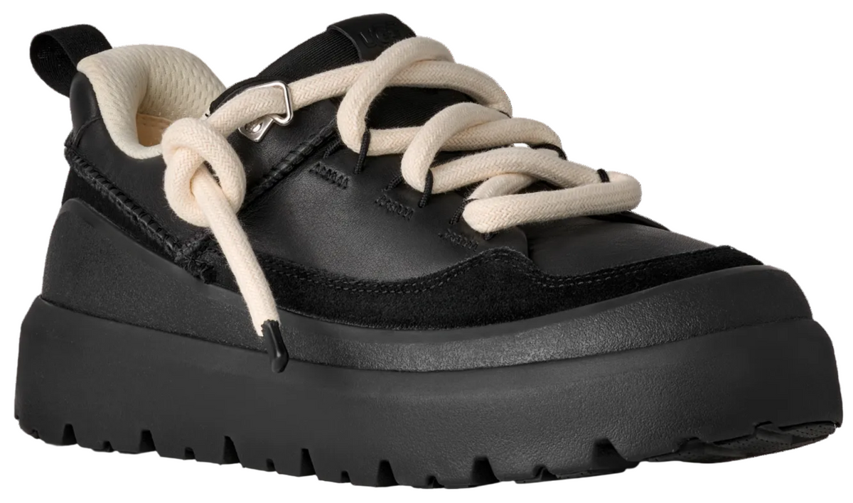 A black low-top platform sneaker with a thick, jagged-edged rubber sole, featuring thick cream-colored, knotted laces and a cream-colored inner lining.
