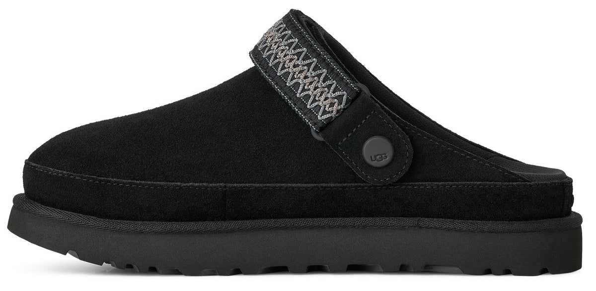 A black suede mule slip-on shoe with a thick sole and a decorative woven strap across the instep featuring a small circular branded button on the side.
