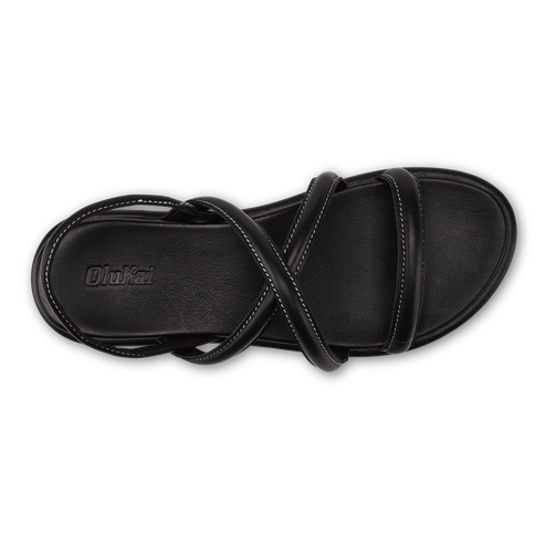 A top-down view of a black women's flat sandal with a cushioned footbed, featuring crisscrossing thin straps across the instep and an ankle strap with visible stitching and the embossed brand name OluKai on the heel.