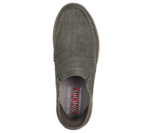 Top-down view of a green canvas slip-on shoe, showcasing visible stitching on the sides and the insole labeled "SLIP-INS with exclusive HEEL PILLOW".