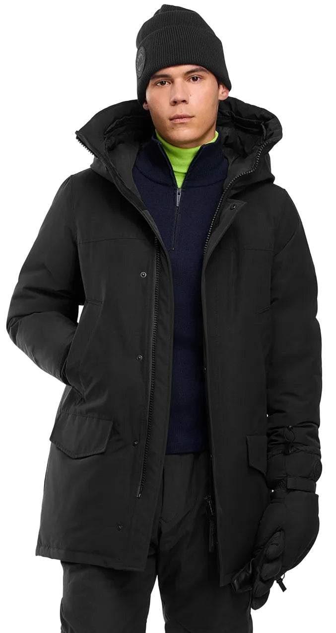 A male model wearing a black winter parka with a large hood, a navy blue quarter-zip fleece over a neon green mock-neck shirt, black gloves, and a black beanie.
