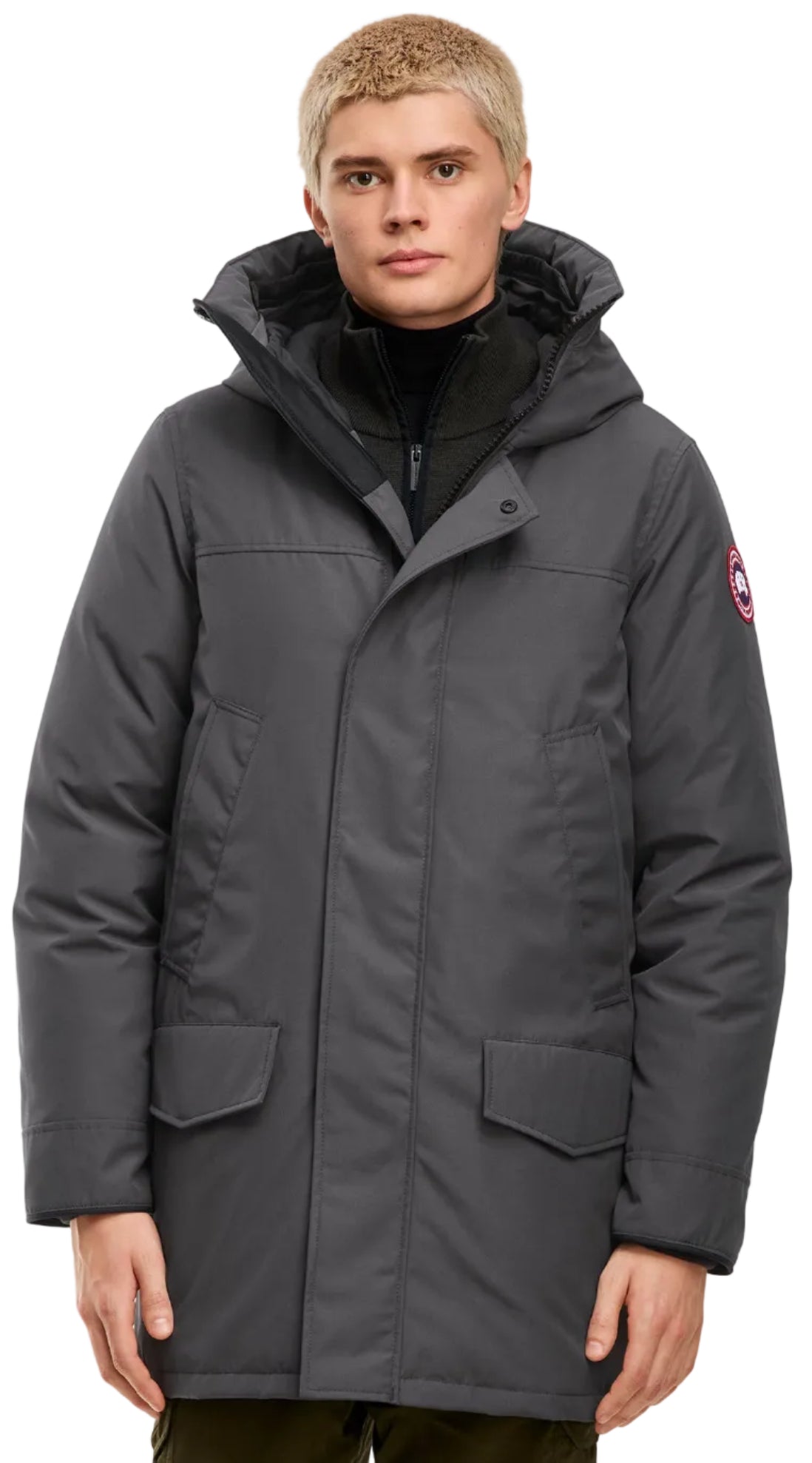 A fair-skinned man with short blonde hair wears a charcoal gray, hooded, mid-length parka featuring a front zipper hidden by a buttoned storm flap, two large front flap pockets, and a circular red, white, and blue logo patch on the left sleeve.