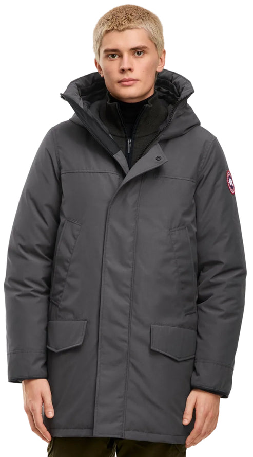 A fair-skinned man with short blonde hair wears a charcoal gray, hooded, mid-length parka featuring a front zipper hidden by a buttoned storm flap, two large front flap pockets, and a circular red, white, and blue logo patch on the left sleeve.