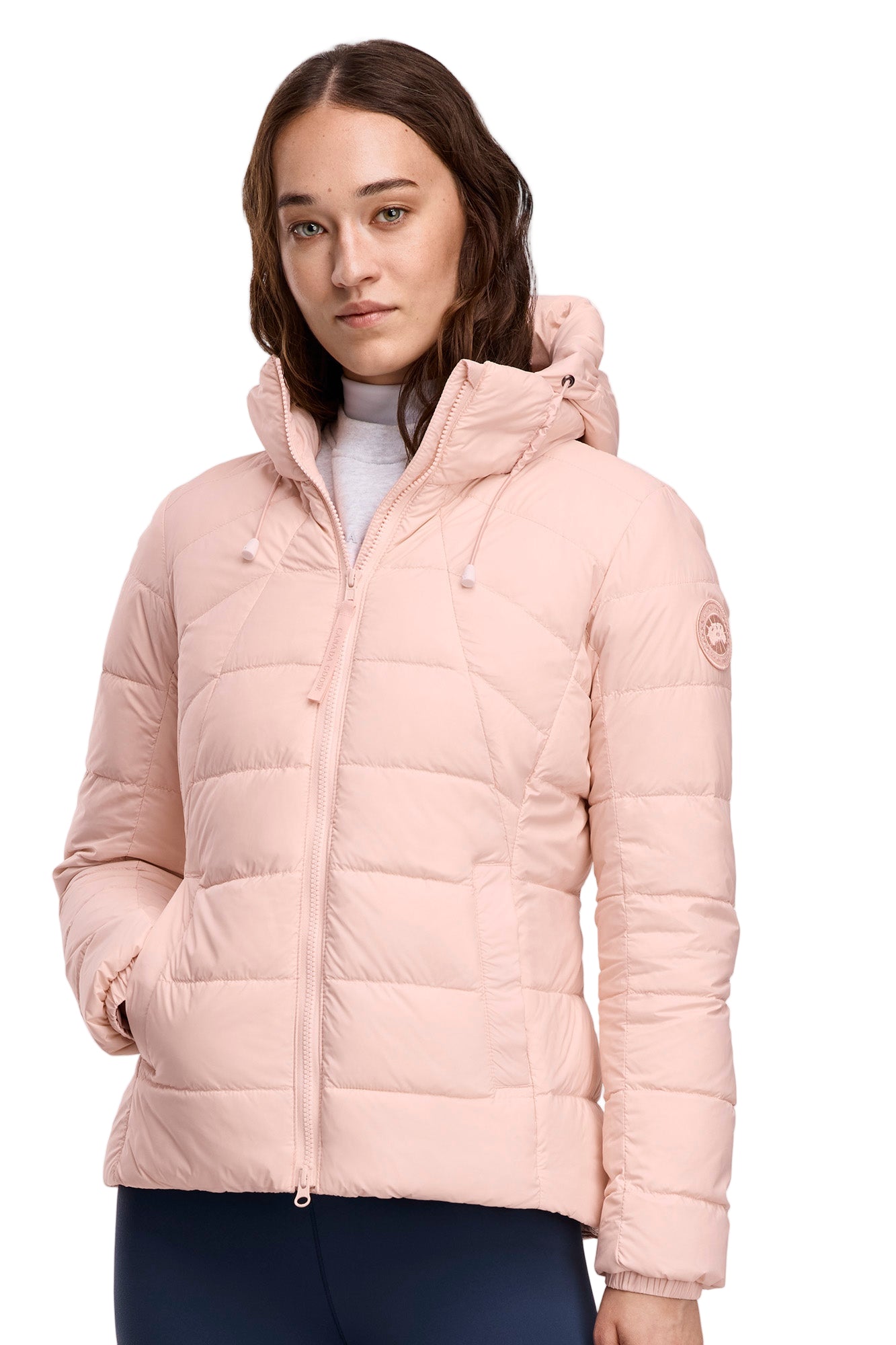 A woman wearing a light pink hooded, down-filled puffer jacket with a front zipper and an emblem on the left shoulder.