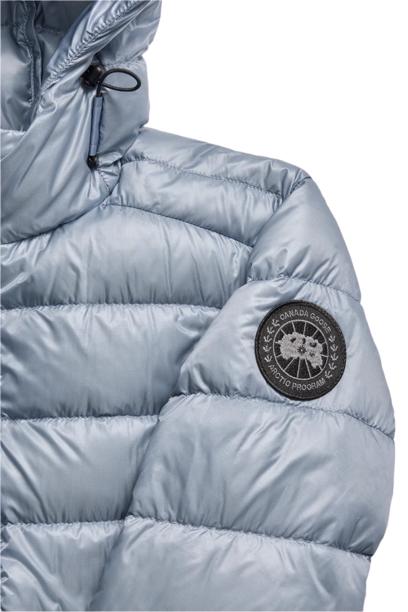 Close-up of a shiny, metallic light blue quilted puffer jacket with a hood, featuring a round, black logo patch on the upper sleeve.