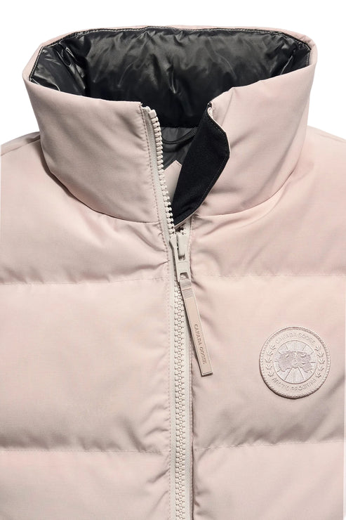 A close-up view of a pale pink, quilted down vest with a high collar featuring a black interior lining, a central zipper with a matching pull tab, and a circular, monochrome logo patch on the right chest.