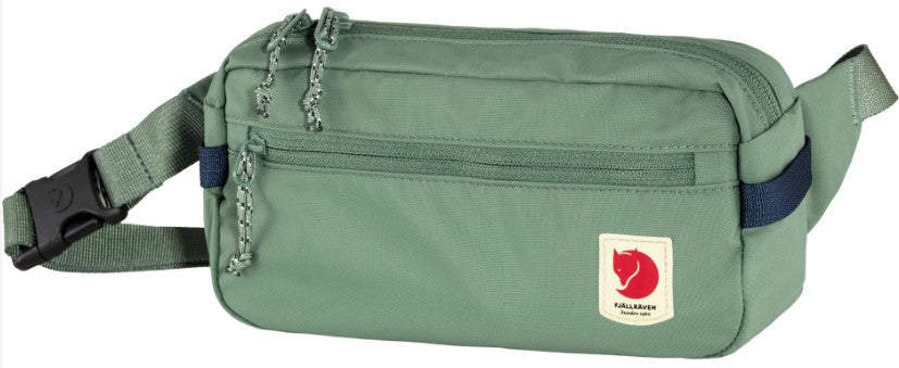A sage green waist pack with a horizontal zippered pocket across the front, a main zippered compartment with dual pulls, and a dark blue strap accent on the side. A small off-white patch with a red fox logo and the text Fjällräven is stitched on the lower right corner.