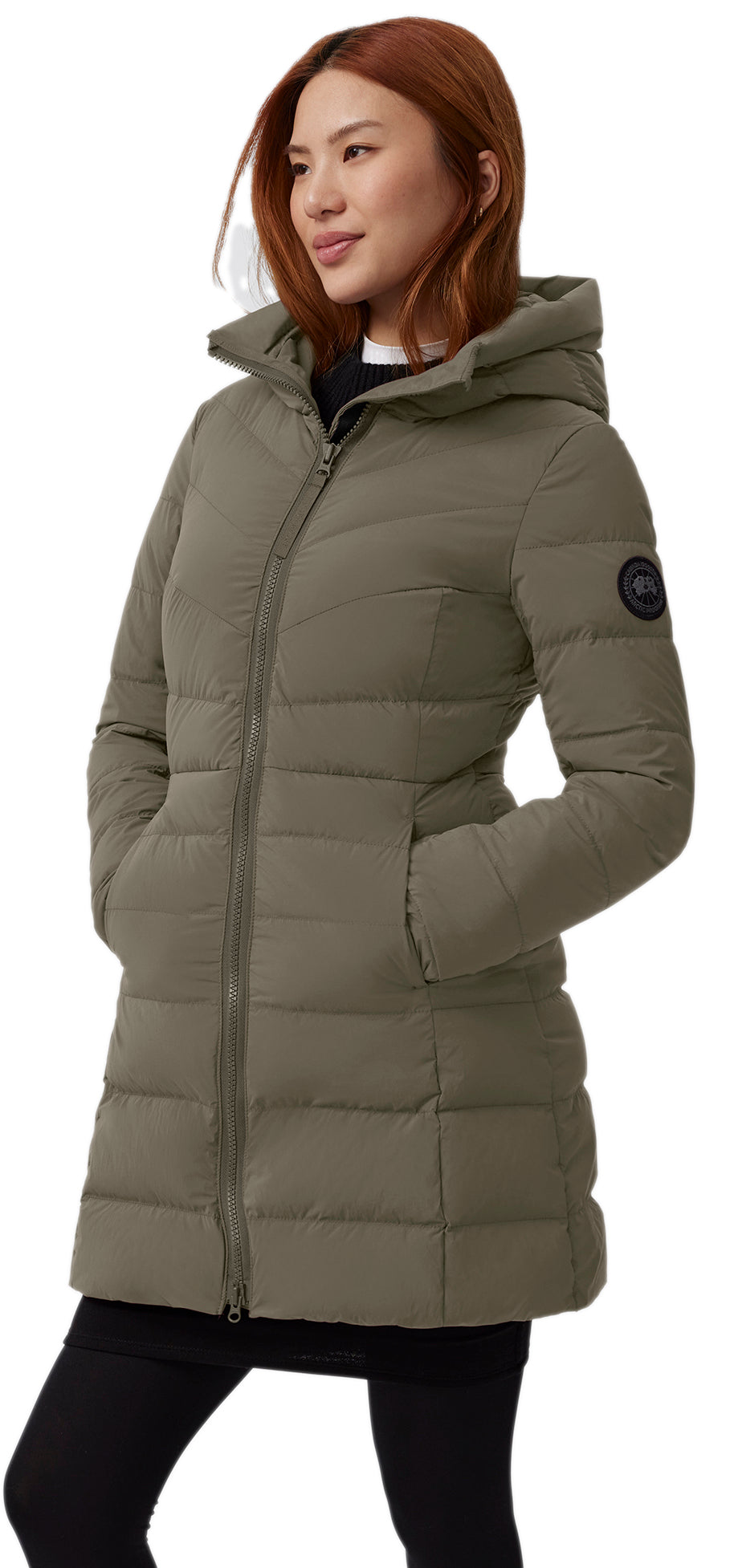 A woman wearing a padded olive coat with a hood and a black circular logo on the arm.