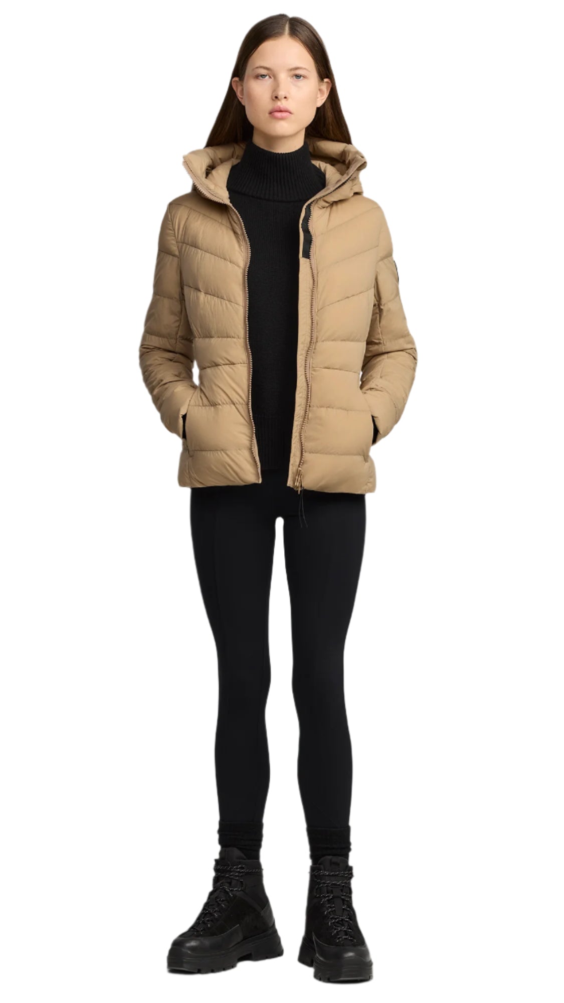 A person wearing a tan puffer jacket with a hood, black turtleneck sweater, black leggings, and black boots.