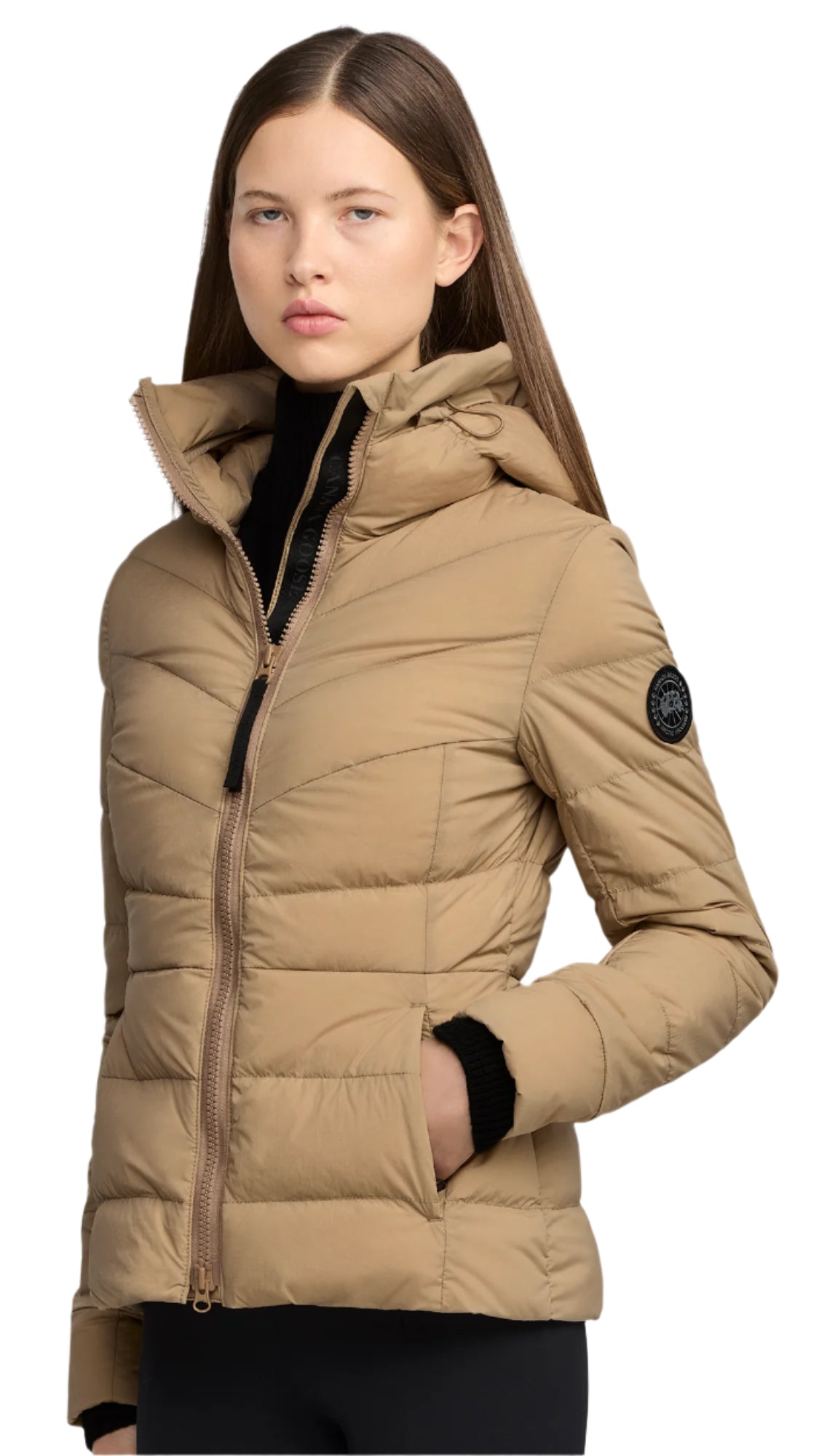 A woman wearing a tan, quilted jacket with a hood and a black patch on the upper arm.