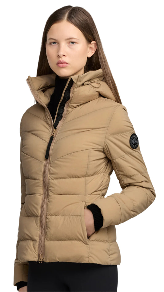 A woman wearing a tan, quilted jacket with a hood and a black patch on the upper arm.