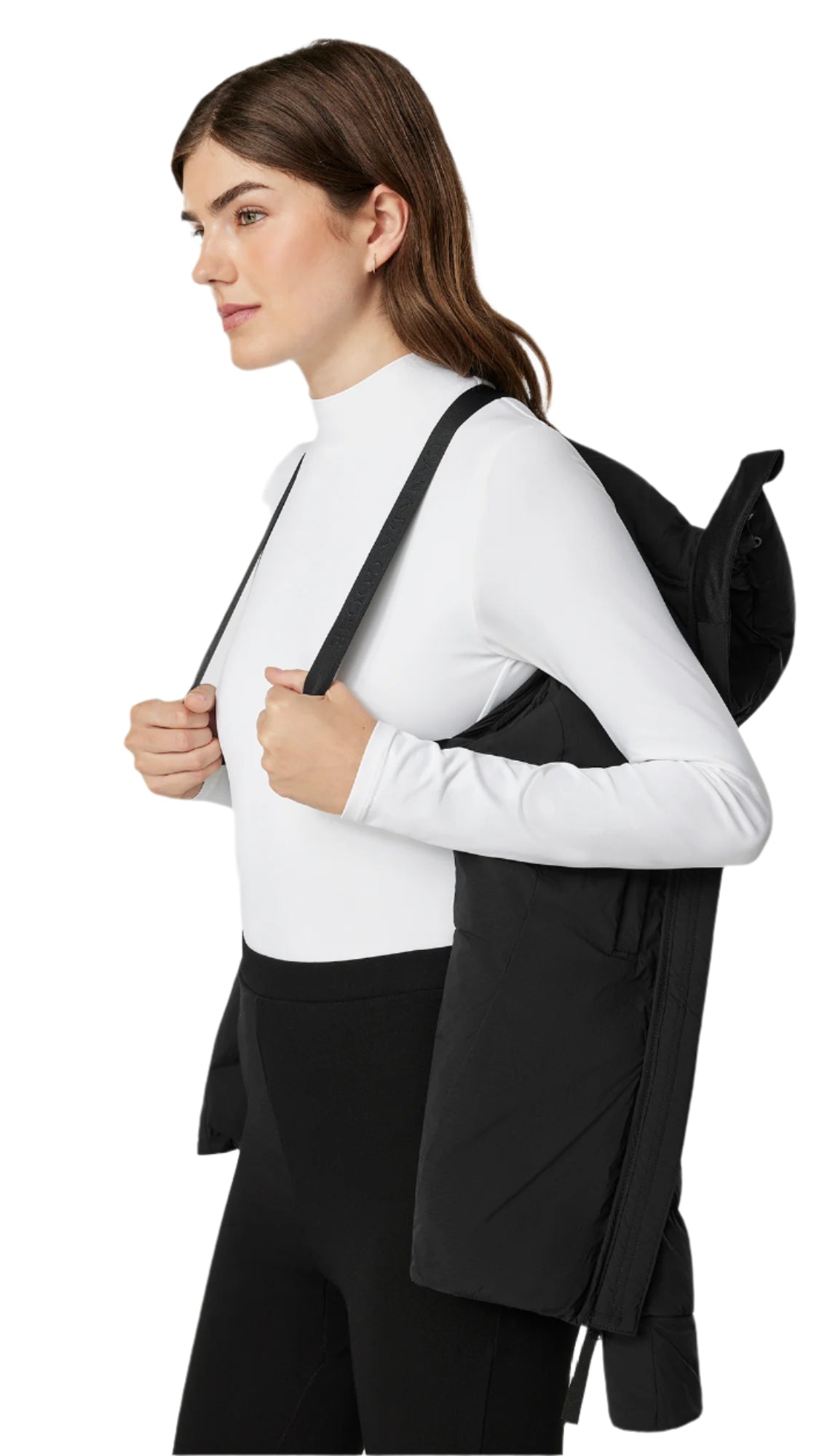 A woman wearing a white turtleneck and black leggings is standing sideways, wearing a black jacket hanging on her back with straps.