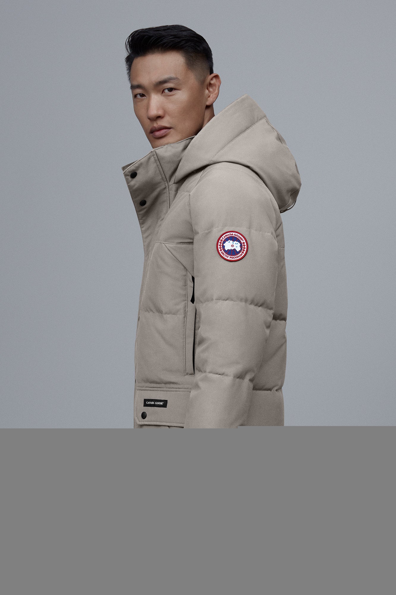 A person is wearing a tan puffer coat with a hood and a circular patch with a maple leaf on the left arm.