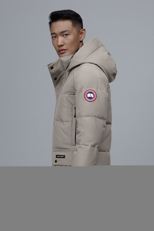 A person is wearing a tan puffer coat with a hood and a circular patch with a maple leaf on the left arm.