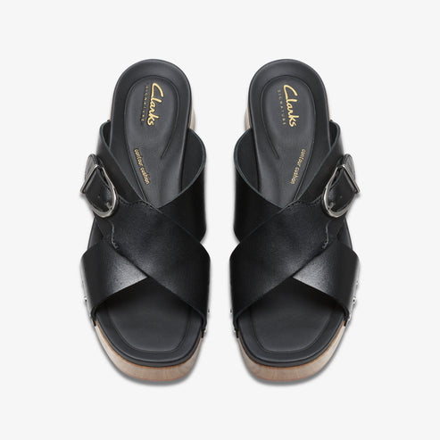 Overhead view of two black leather slide sandals with buckle, on a wooden sole. "Clarks signature" text is visible on the inner sole.