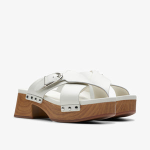 A pair of white, platform sandals with thick straps and a silver buckle, on a light brown, wood-grain sole.