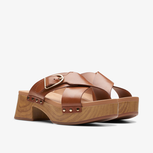 Tan leather sandal with crisscrossing straps and gold buckle on a wooden platform heel.