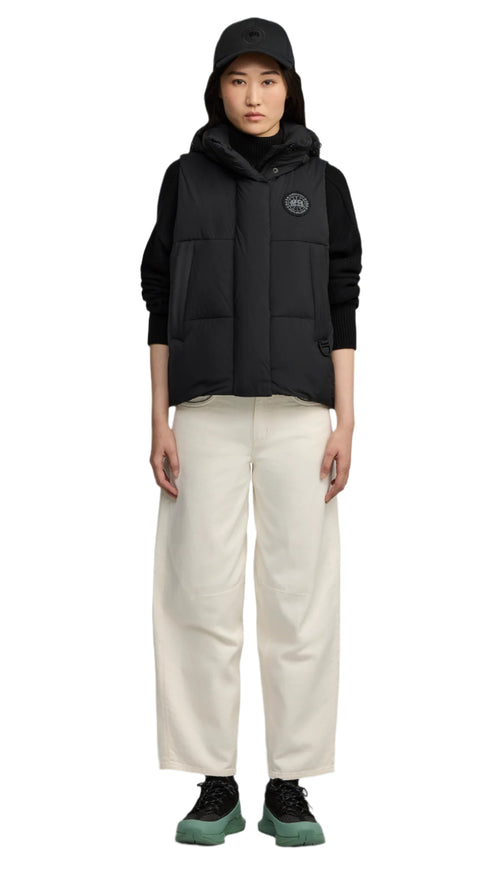 A woman wearing a black vest, black shirt, white pants, black shoes and a black hat.