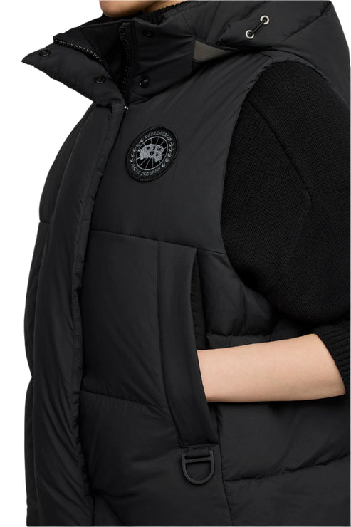 A black puffer vest with a hood and black circular logo emblem on the upper left side, and a hand in the side pocket.