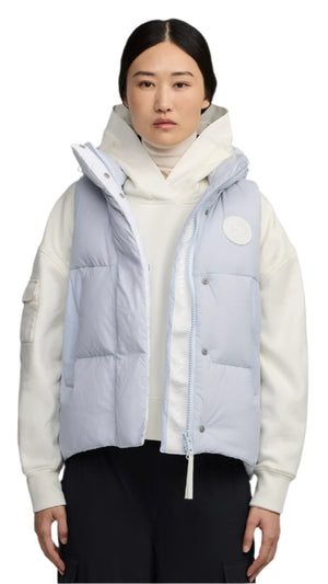 A woman wears a light blue puffy vest over a white sweatshirt with black pants.